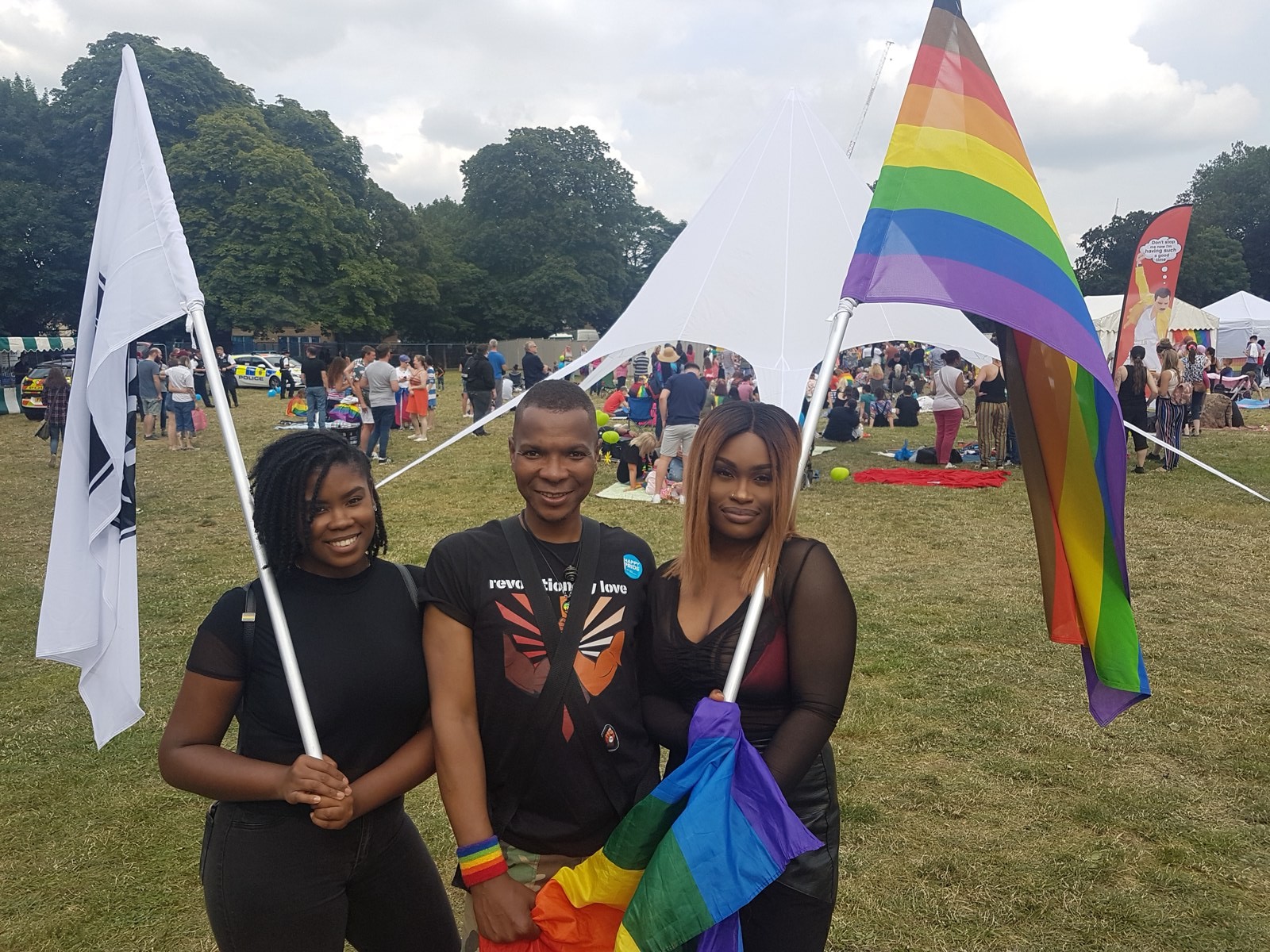 Three smiling young people holding flags at the BLKOUT stand at Croydon Pride, one in a revolutionary love t-shirt