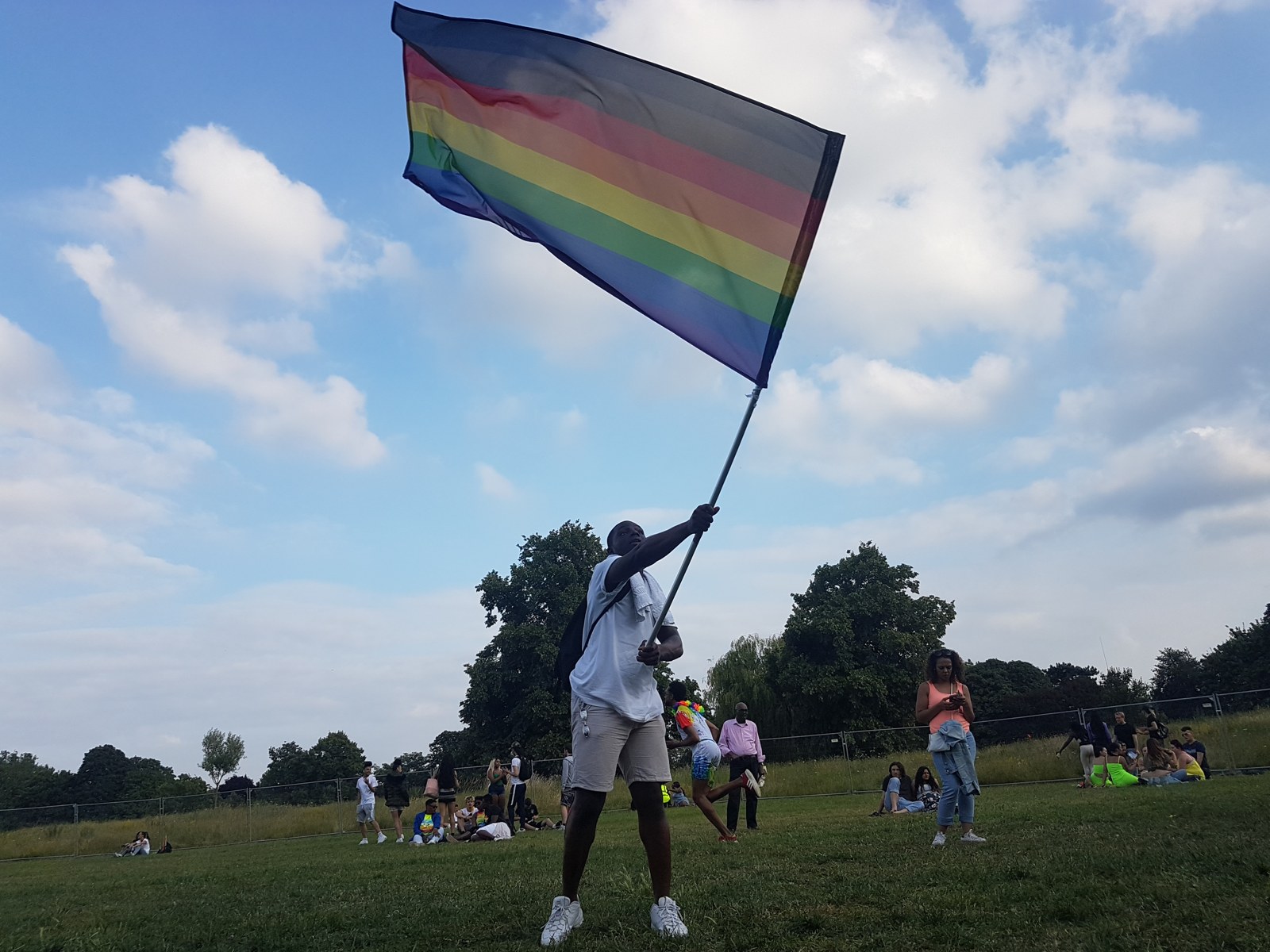 A young Black man waves a huge Philadelphia-stripes rainbow flag against the sky in a Croydon park
