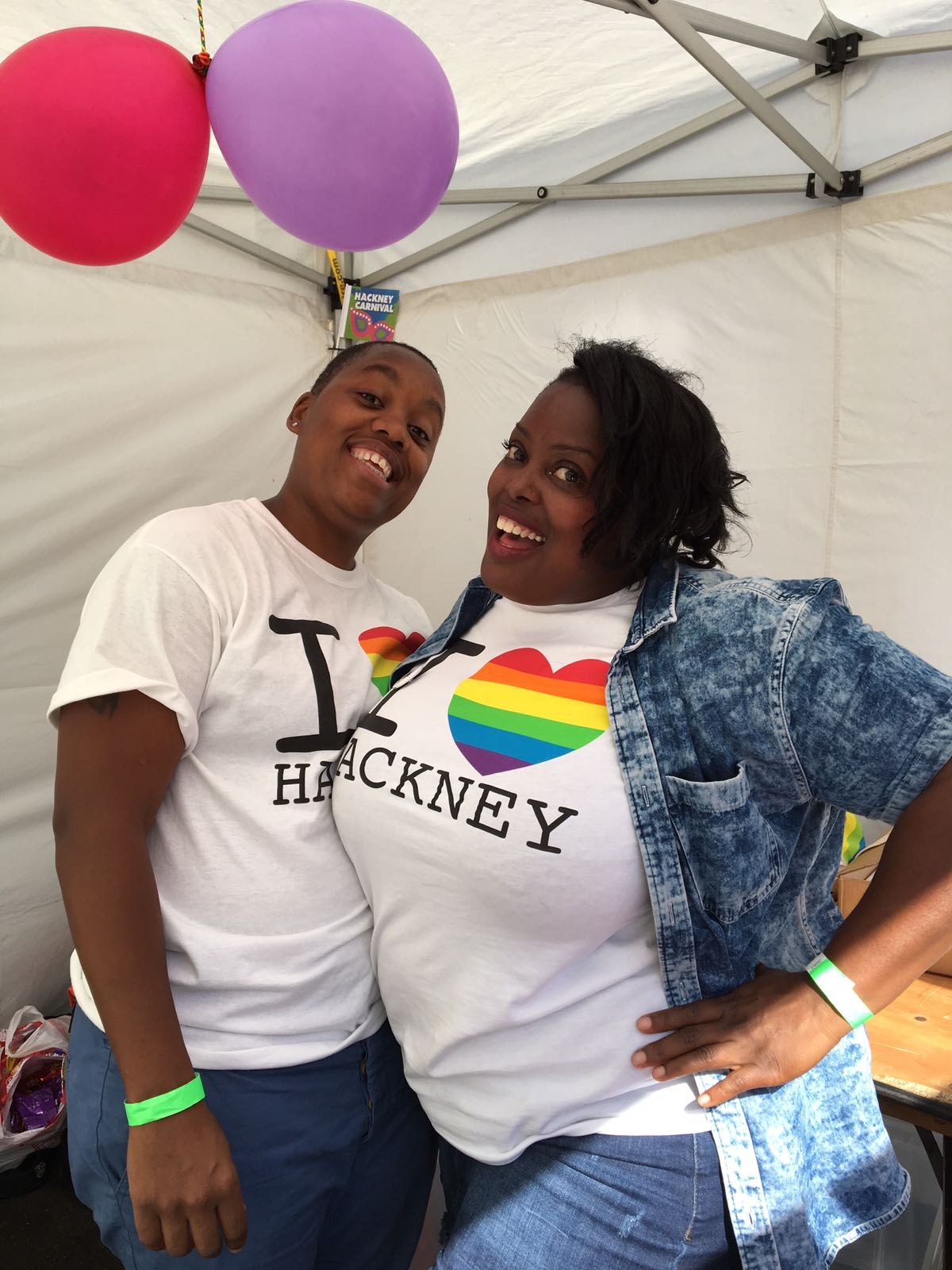 Two women smiling closely together, both wearing white T-shirts printed with a rainbow heart and the words I LOVE HACKNEY, under a gazebo with balloons
