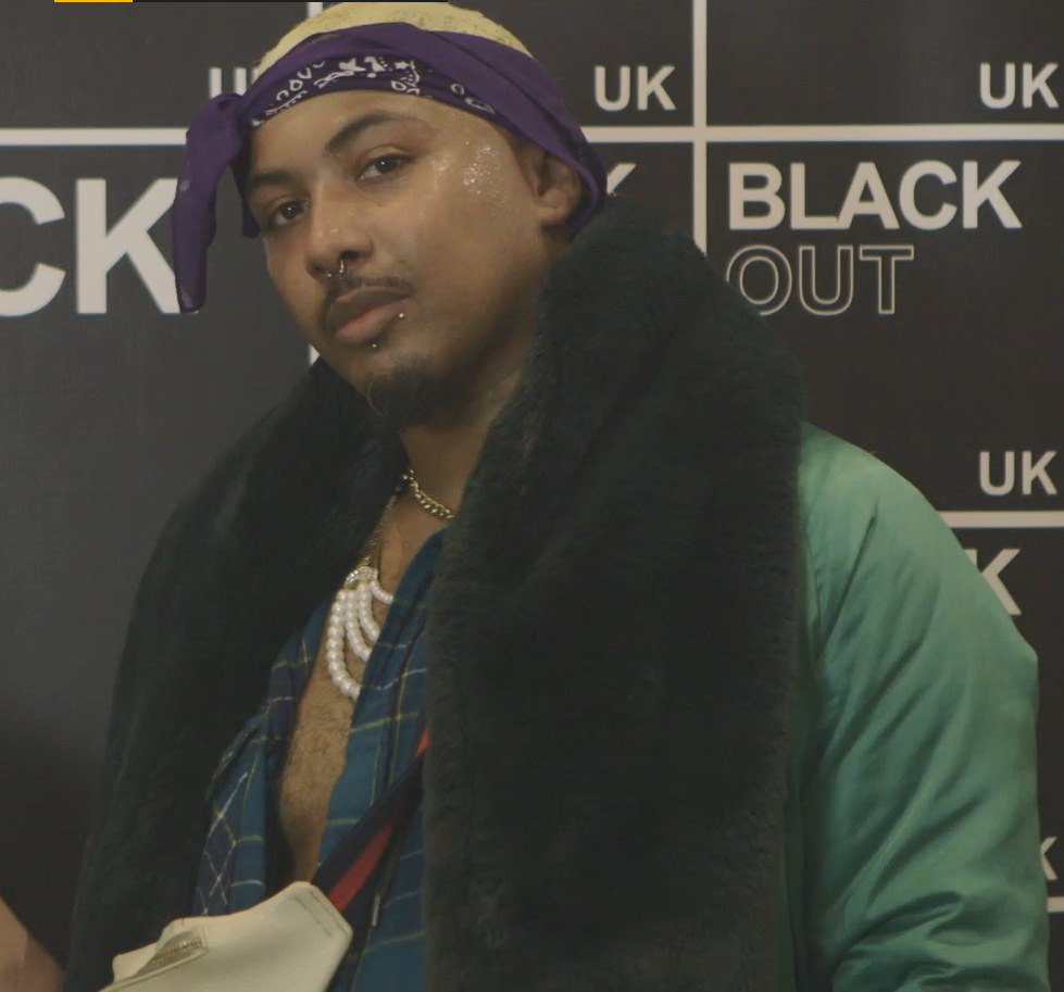 A man in a purple bandana, dark fur stole and pearls poses at the BLACK OUT UK wall